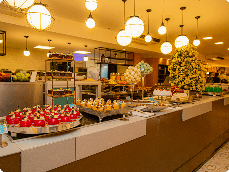 Buffet de natal, com uma grande variedades de doces a vista no Restaurante Taboa. Pendentes de luz mais amarelados presos no teto. No funda há algumas frutas, opções de sucos.