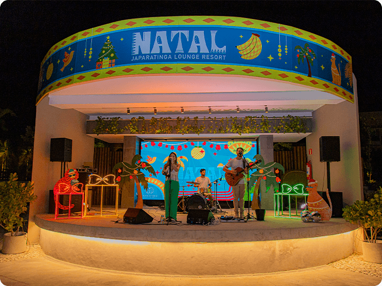 Palco do Natal da Praça dos Coqueiros no Japaratinga Lounge Resort. Uma decoração natalina em cima do palco, com uma banda de 3 integrantes, 2 vocalista sendo um homem e uma mulher e um na bateria. Há luzes coloridas no palco todo e coqueiros ao redor.