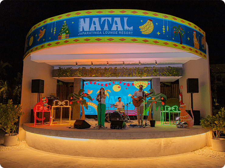 Palco do Natal da Praça dos Coqueiros no Japaratinga Lounge Resort. Uma decoração natalina em cima do palco, com uma banda de 3 integrantes, 2 vocalista sendo um homem e uma mulher e um na bateria. Há luzes coloridas no palco todo e coqueiros ao redor.