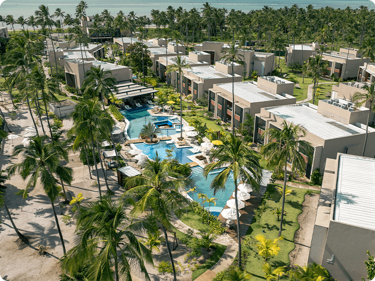 Vista aérea das Gardens Suítes do Japaratinga Lounge Resort, mostrando a área das piscinas com guarda-sóis brancos, espreguiçadeiras, cercadas por vegetação tropical e palmeiras