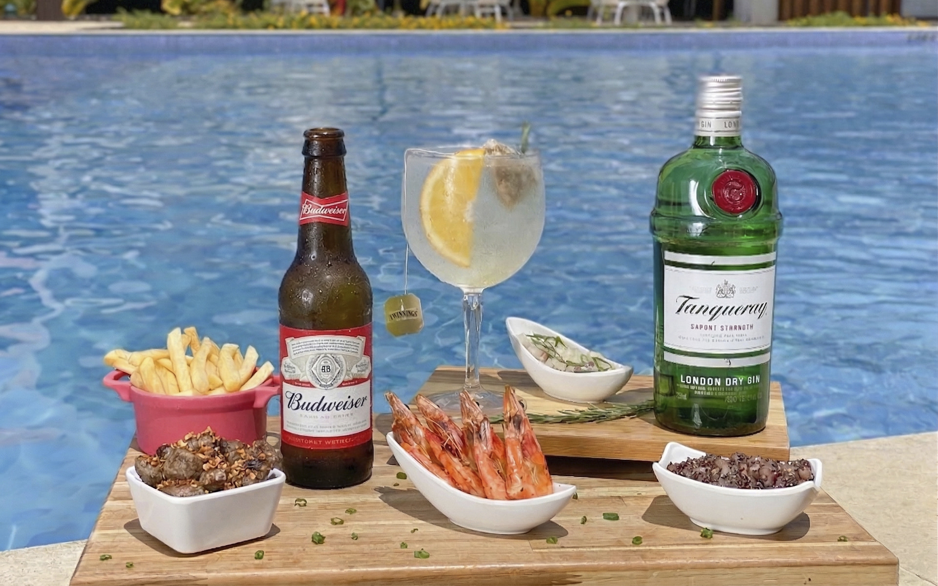 Mesa à beira da piscina do Japaratinga Lounge Resort com gin Tanqueray, cerveja Budweiser, camarões, batata frita, ceviche e petiscos, com área de lazer ao fundo.
