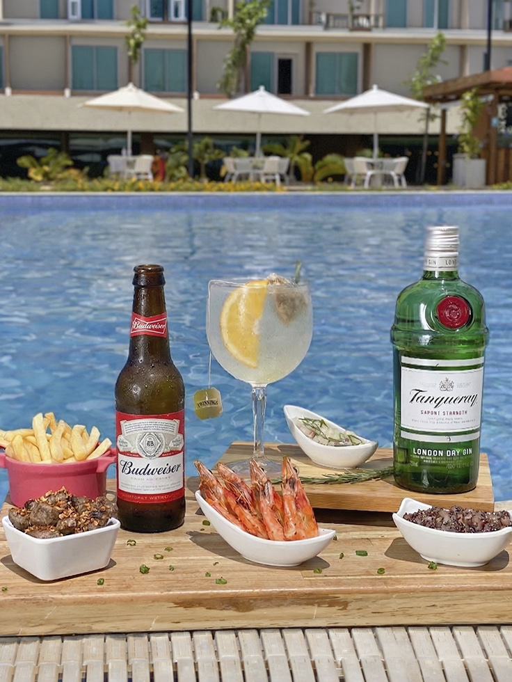 Mesa à beira da piscina do Japaratinga Lounge Resort com gin Tanqueray, cerveja Budweiser, camarões, batata frita, ceviche e petiscos, com área de lazer ao fundo.