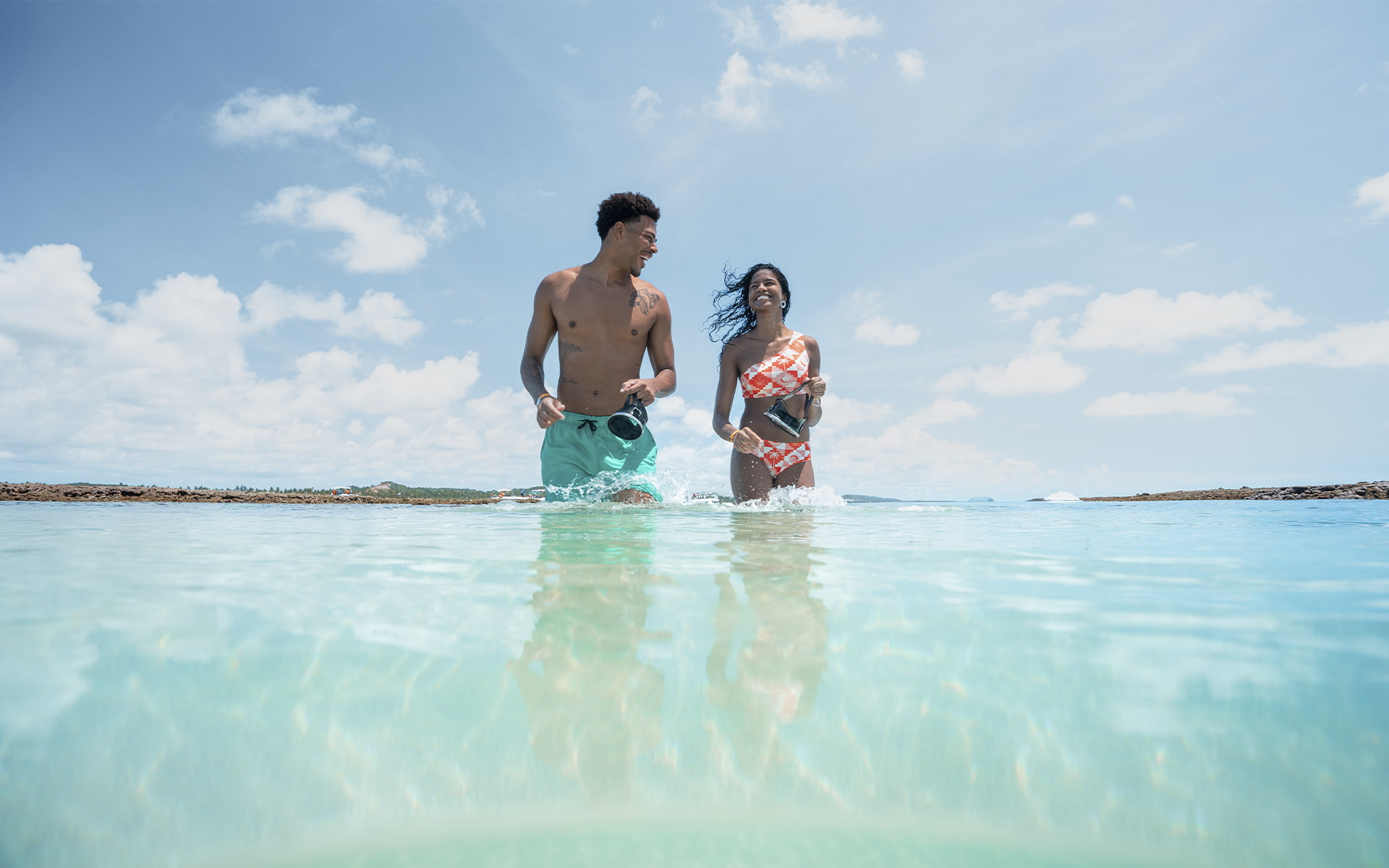 Casal caminhando e sorrindo nas águas cristalinas das piscinas naturais de Japaratinga, com recifes ao fundo em dia de céu azul.