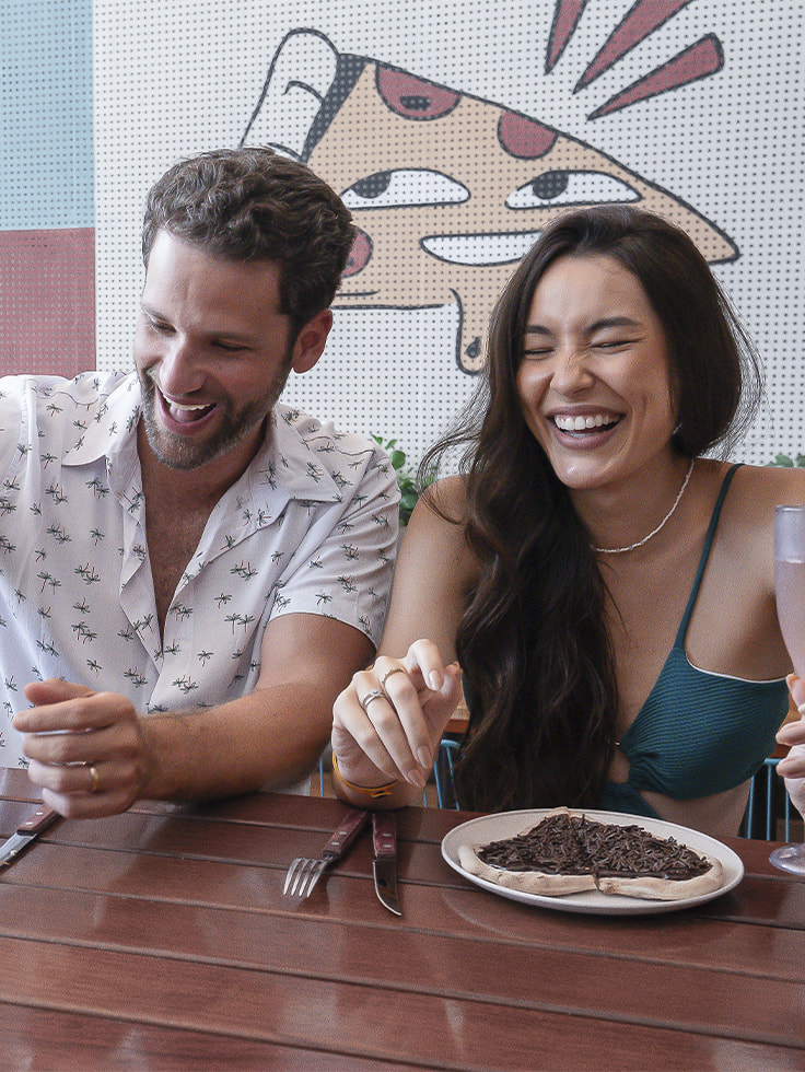 Casal sorrindo à mesa da Amore Forneria com pizzas artesanais, cerveja Corona e taça de espumante, cercado por parede com arte colorida.