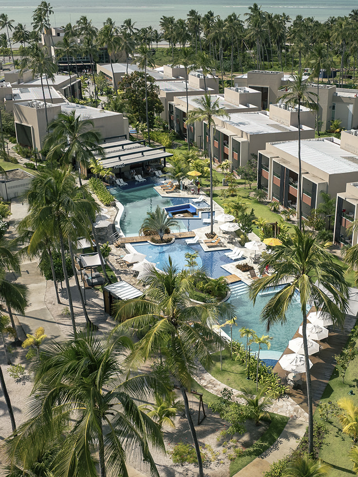 Vista aérea da área Garden do Japaratinga Lounge Resort com complexo de piscinas conectadas e vista para o Mangô Bar, cercado por estrutura moderna de quartos e extenso coqueiral, com o mar ao fundo.