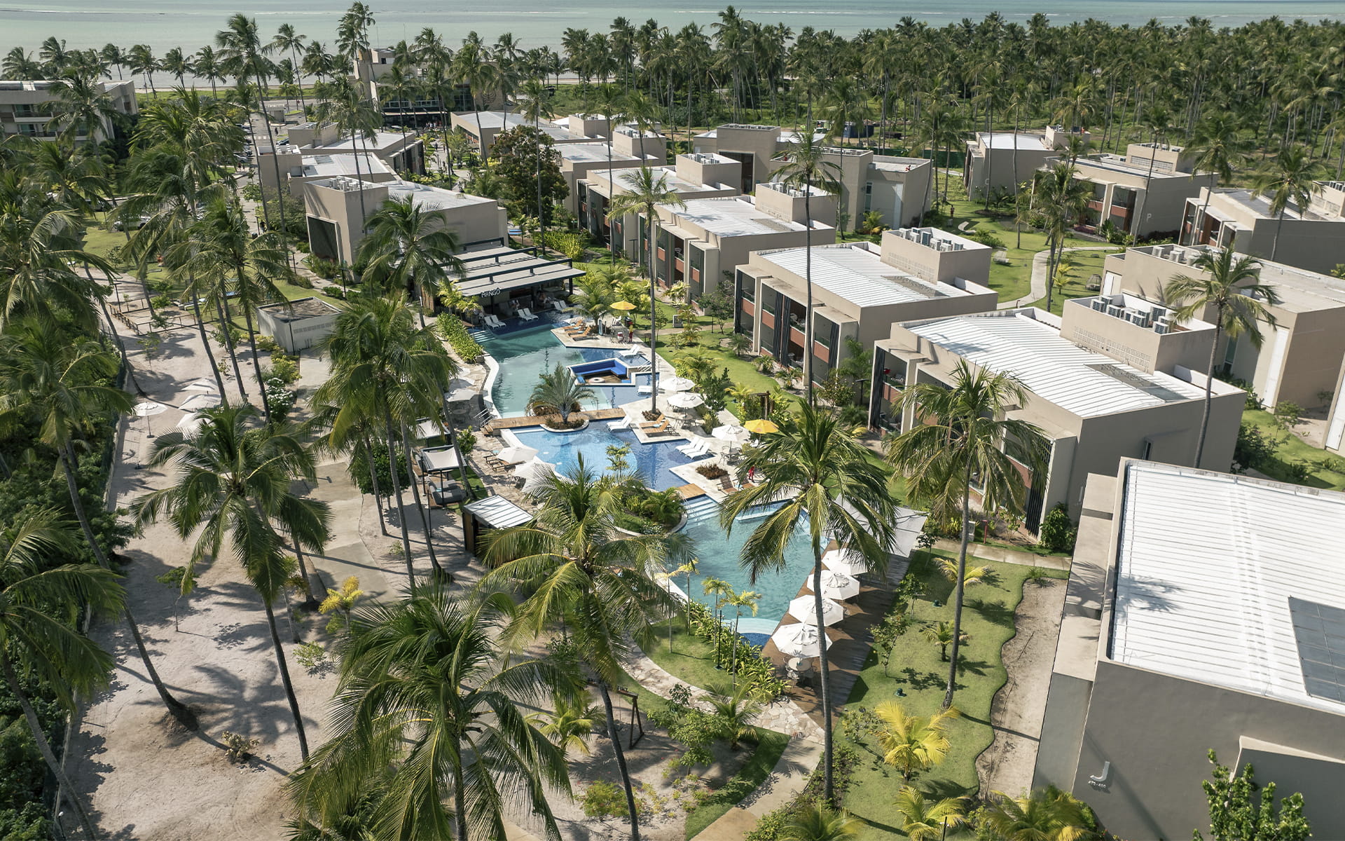 Vista aérea da área Garden do Japaratinga Lounge Resort com complexo de piscinas conectadas e vista para o Mangô Bar, cercado por estrutura moderna de quartos e extenso coqueiral, com o mar ao fundo.