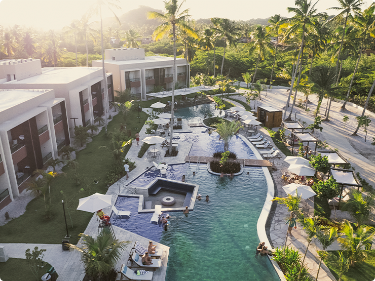 Uma vista aérea da piscina rodeado por coqueirais. Ao redor, tem os quartos Gardens, decks, espreguiçadeiras e pessoas aproveitando a água. A luz é intensa e dourada, típica do pôr do sol.