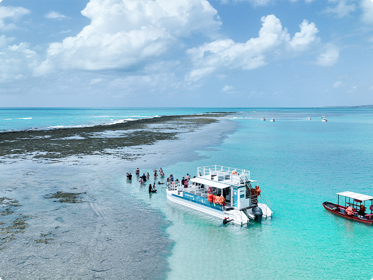 Vista aérea da praia de Japaratinga, com um catamarã em águas cristalinas, famílias estão nas águas das piscinas naturais, proporcionando um cenário paradisíaco.
