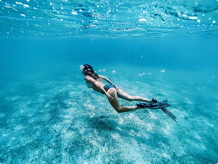 Mulher com pé de pato e óculos de mergulho, nadando em águas cristalinas e azuis. A luz do sol reflete na água que é possível enxergar o chão.