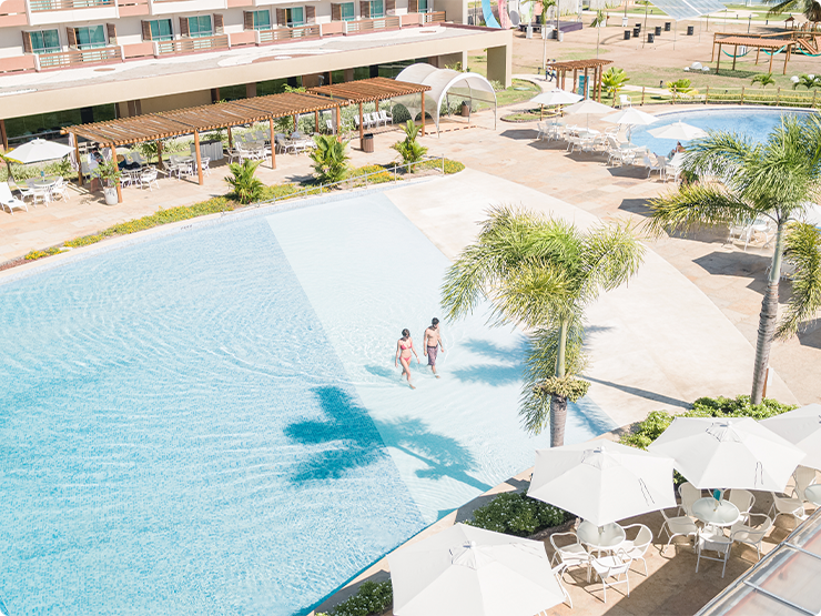 Vista aérea de uma grande área de piscina do Japaratinga Lounge Resort. O local possui guarda-sóis brancos, espreguiçadeiras e bangalôs de madeira. Um casal caminha na parte rasa da piscina. A estrutura do resort é visível ao fundo.