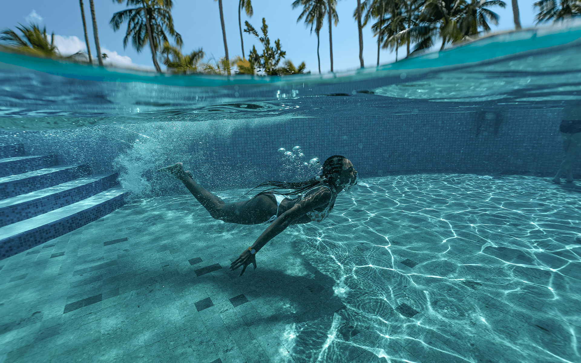 Mulher mergulhando na piscina das Gardens do Japaratinga Lounge Resort. Acima é possível ver os coqueiros em foto subaquática.