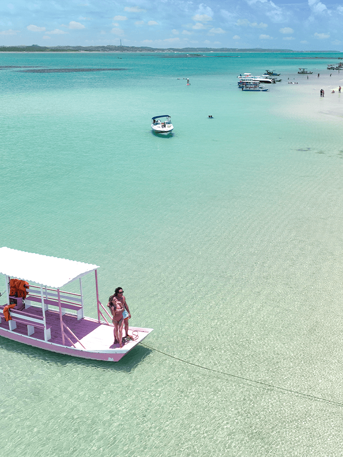 Passeio de jangada ancorada em águas cristalinas das piscinas naturais da praia de Japaratinga. Possível ver os recifes de corais e casal em cima da jangada rosa.