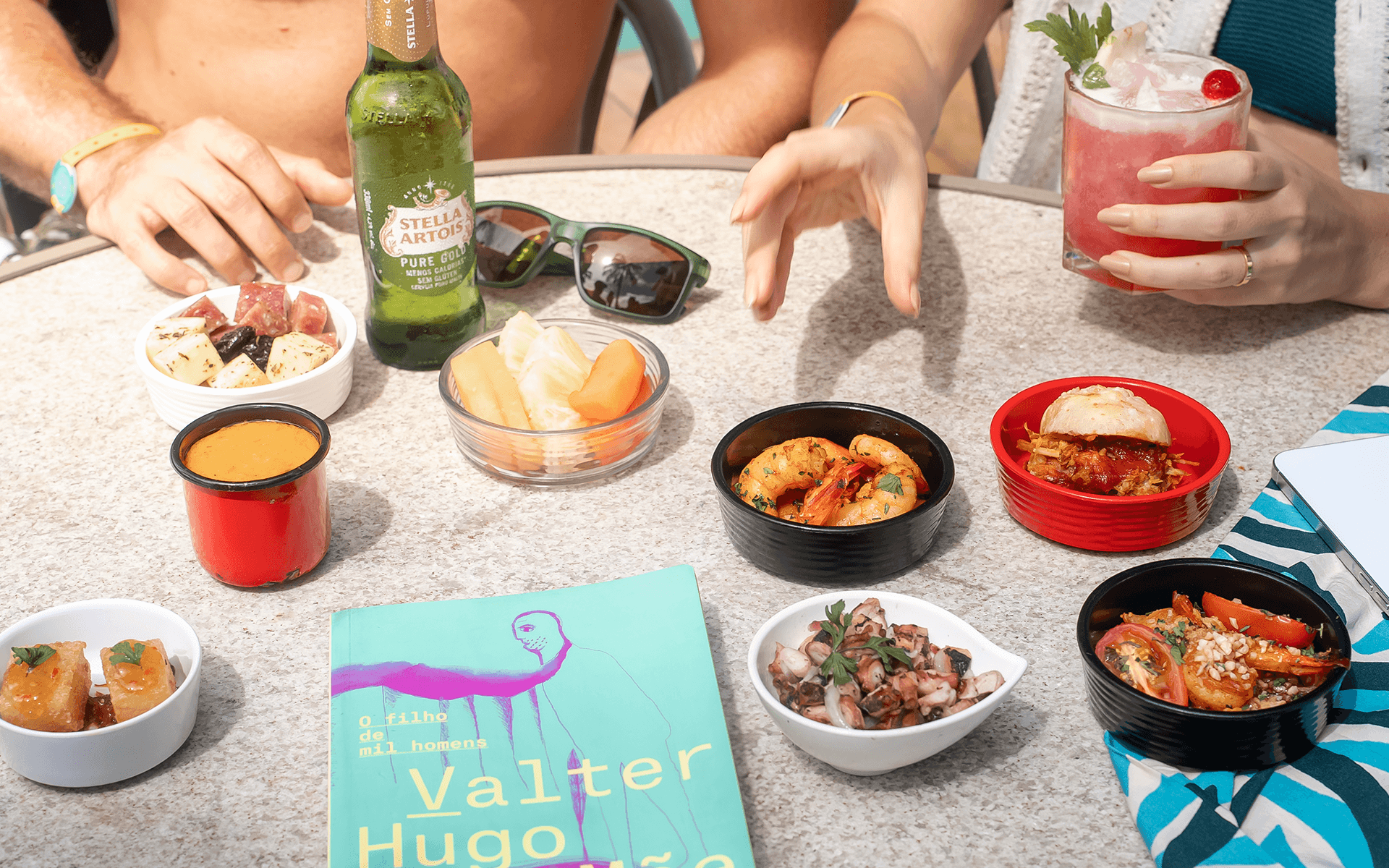 Mesa do All Inclusive Premium com pratos e petiscos, queijos, camarões hamburguer e bebidas como Cerveja Stella Artois e drink servidos com casal na mesa.