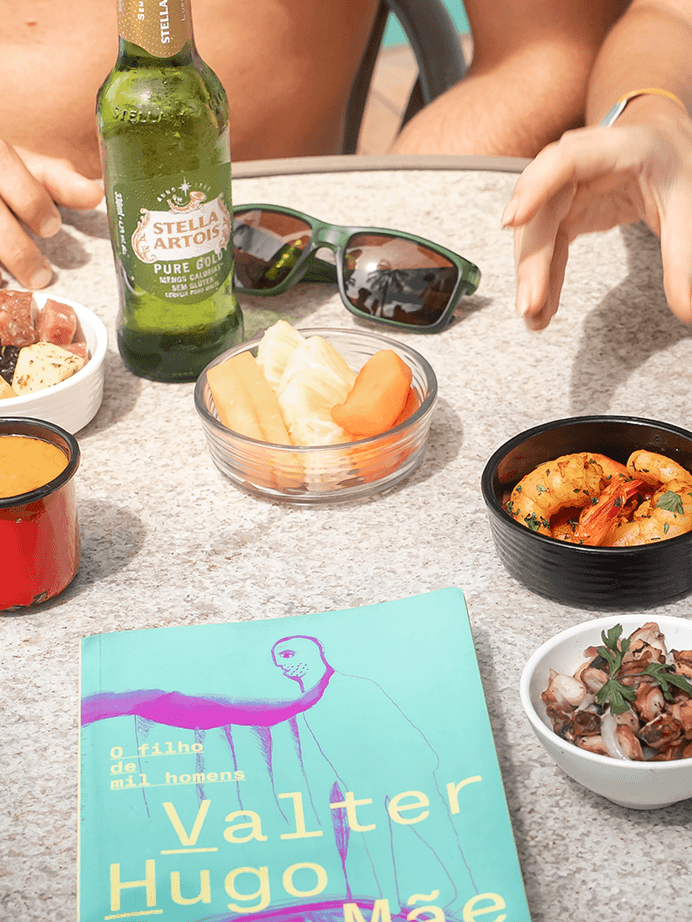 Mesa do All Inclusive Premium com pratos e petiscos, queijos, camarões hamburguer e bebidas como Cerveja Stella Artois e drink servidos com casal na mesa.