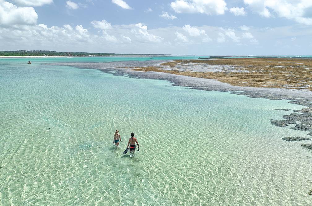 Casal caminhando em águas cristalinas nas piscinas naturais da praia de São miguel dos milagres. É possível ver de cima os recifes de corais expostos por esta com maré baixa.