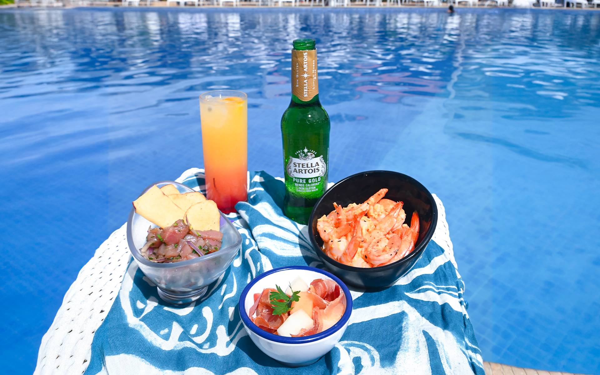 Petiscos de frutos do mar, ceviche, coquetel tropical e cerveja Stella Artois Pure Gold servidos à beira da piscina azul no Japaratinga Lounge Resort.