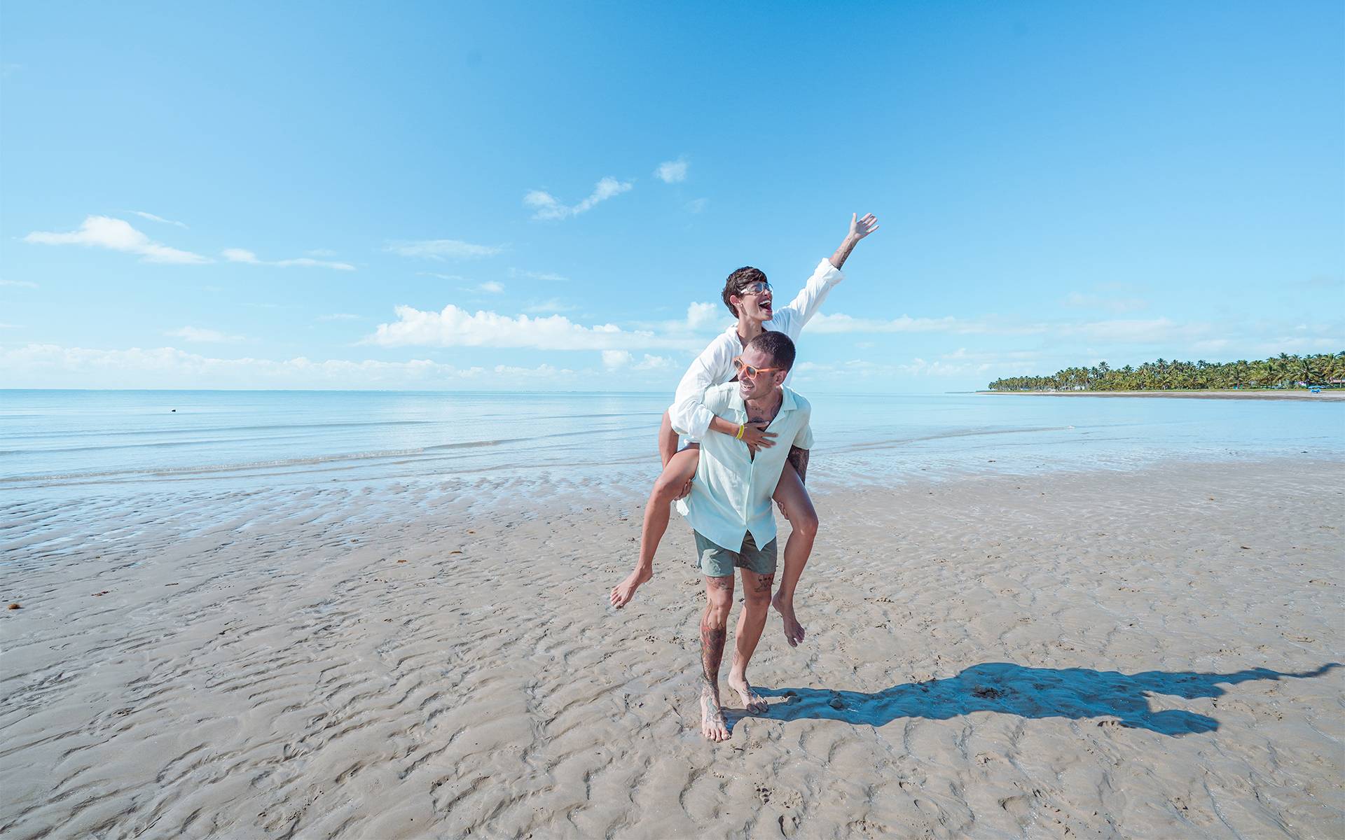 Casal brincando de cavalinho na praia de Japaratinga com mar calmo e areia clara no Japaratinga Lounge Resort, e céu com poucas nuvens.