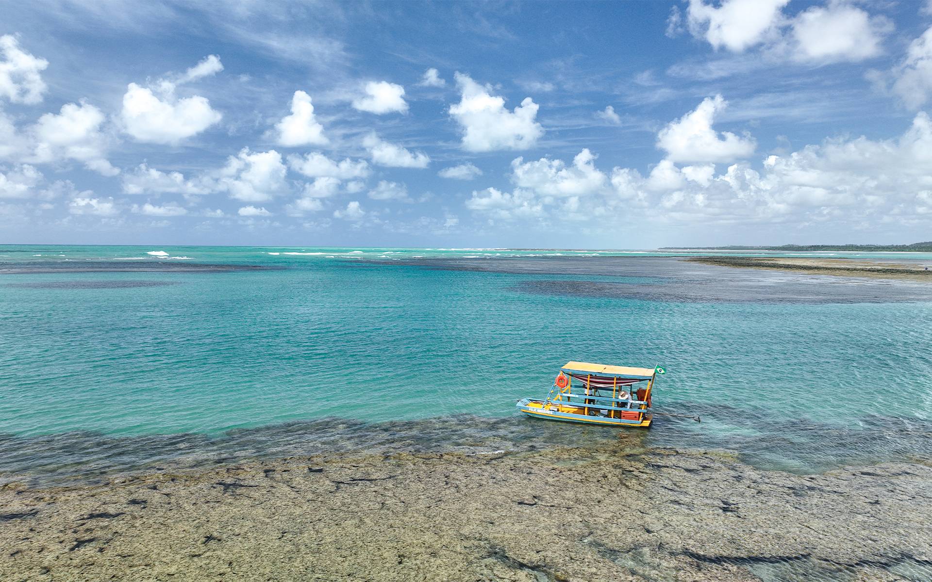 Barco de passeio nas águas azul-turquesa da Praia do Patacho, com recifes de corais visíveis com horizonte do mar ao fundo.