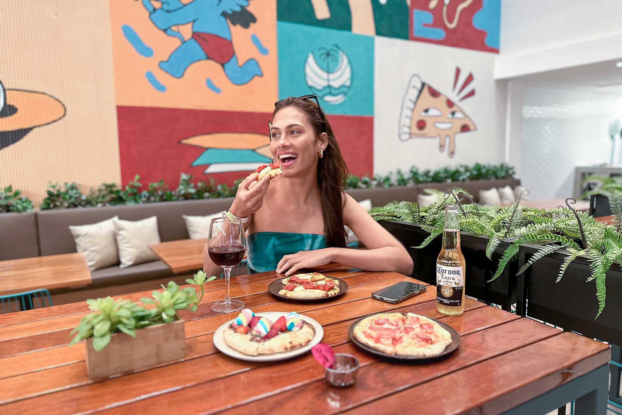 A foto apresenta uma mulher sorridente sentada à mesa em um restaurante aconchegante. Ela está saboreando uma fatia de pizza, com uma taça de vinho tinto ao lado e uma garrafa de Corona Extra à sua frente. O ambiente é decorado com murais coloridos ao fundo.