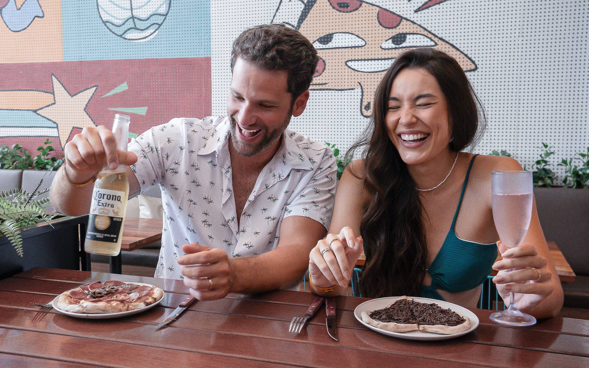 Casal sorrindo no Amore do Japaratinga Lounge Resort, curtindo pizza doce e salgada com cerveja Corona. Um mural com desenhos ao fundo.