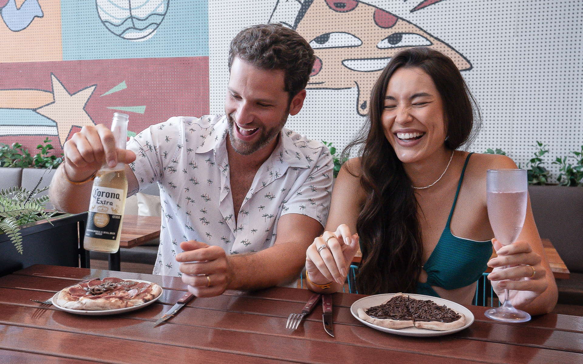 melhor-resort-all-inclusive Casal sorrindo no Amore do Japaratinga Lounge Resort, curtindo pizza doce e salgada com cerveja Corona. Um mural com desenhos ao fundo.