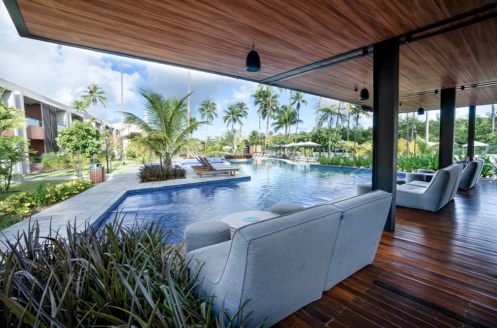 Vista de dentro do Mango Bar em Japaratinga Lounge Resort, na área das Gardens, mostrando puffs pra sentar e piscina, ao redor espreguiçadeiras e muitas plantas.