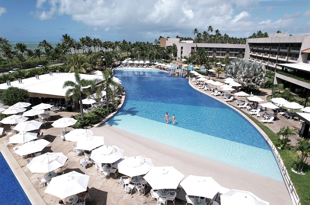 Vista área da piscina principal do Japaratinga Lounge Resort, com Bar Verano a esquerda e estrutura do resort a direita, há inúmeros guarda-sois ao redor da piscina, e muitos coqueiros espalhados.