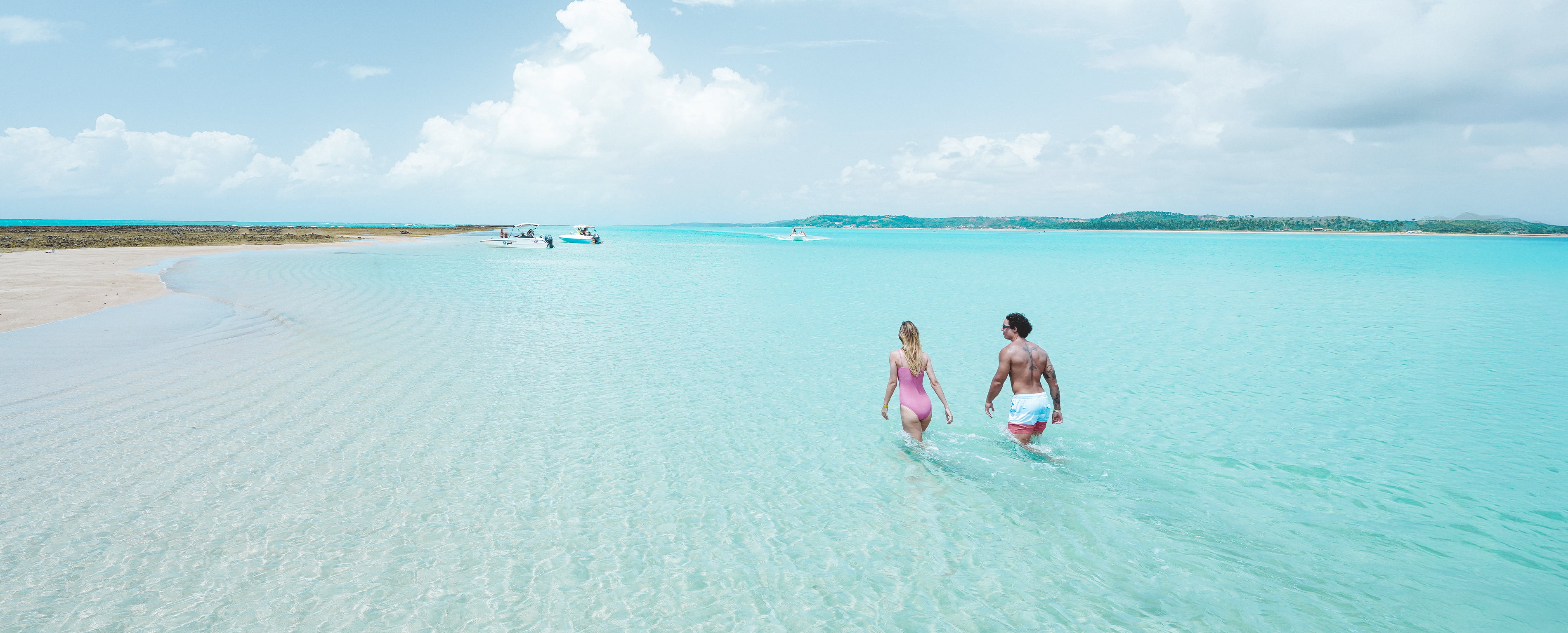 semana-do-consumidor-japaratinga-lounge-resort Casal caminhando na prainha formada pelos recifes de corais e águas cristalinas das piscinas naturais e há um barco ancorado na prainha.