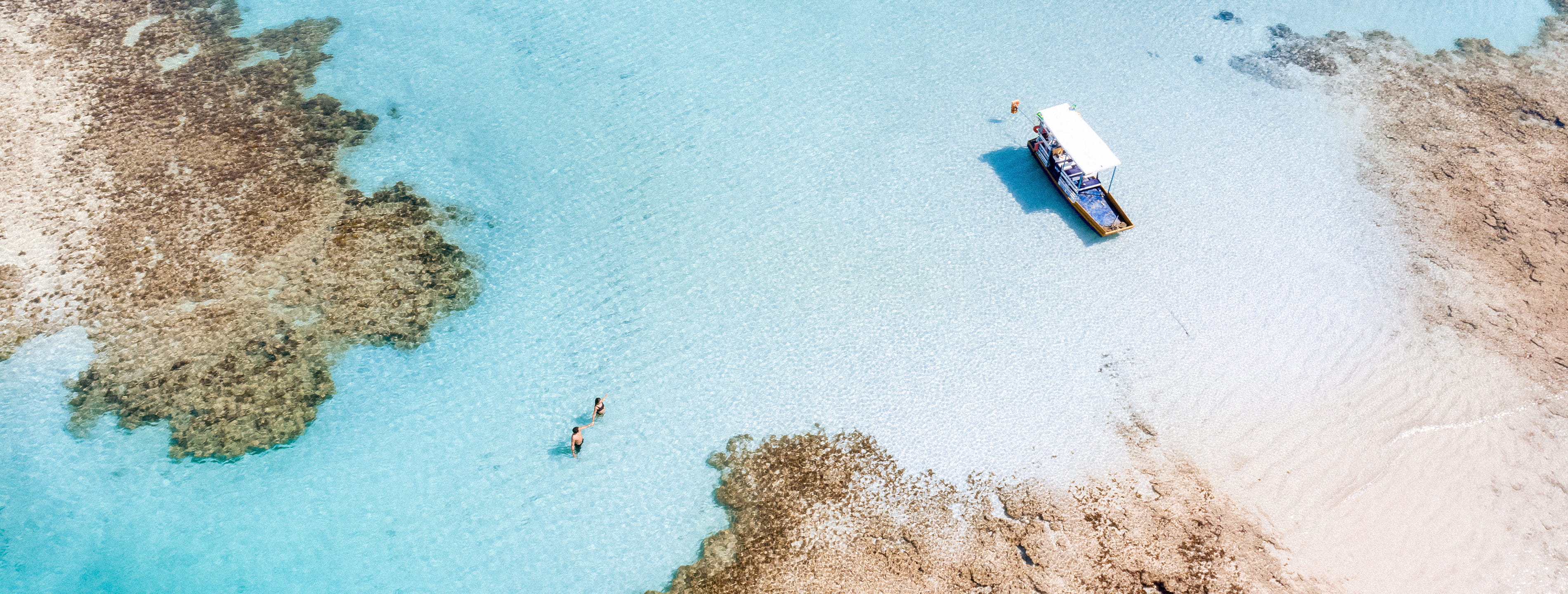 Casal caminhando na prainha formada pelos recifes de corais e águas cristalinas das piscinas naturais e há um barco ancorado na prainha.