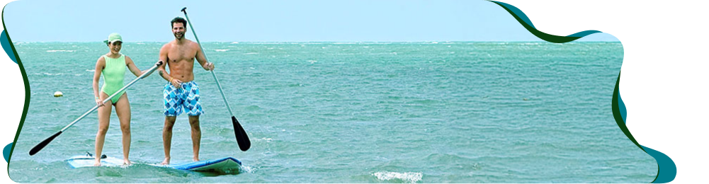 Casal sorrindo e aproveitando as belezas naturais sobre prancha de stand-up padle em meio ao mar da praia de Japaratinga.