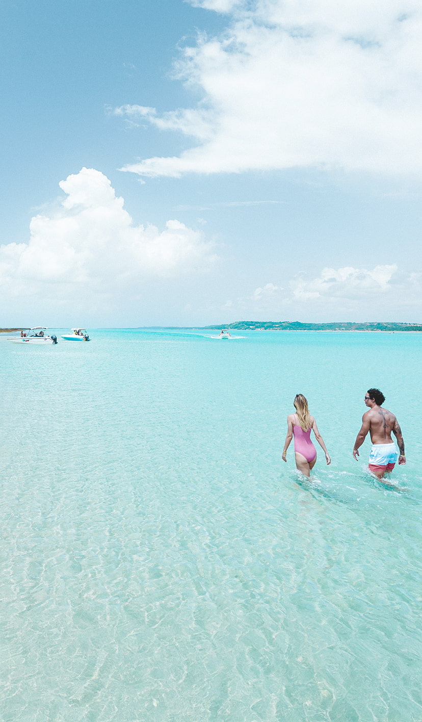 semana-do-consumidor-japaratinga-lounge-resort Casal caminhando na prainha formada pelos recifes de corais e águas cristalinas das piscinas naturais e há um barco ancorado na prainha.