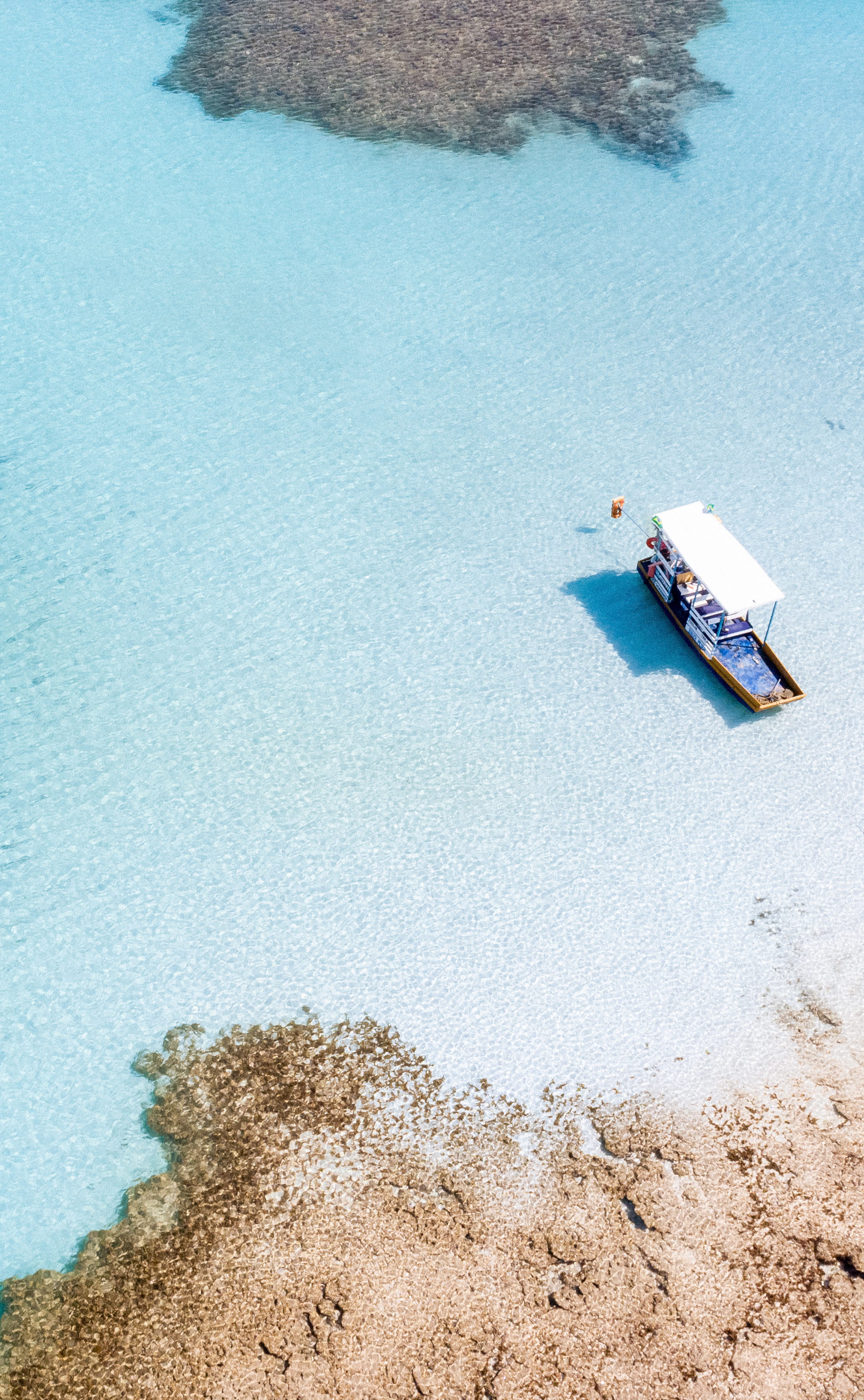 Casal caminhando na prainha formada pelos recifes de corais e águas cristalinas das piscinas naturais e há um barco ancorado na prainha.