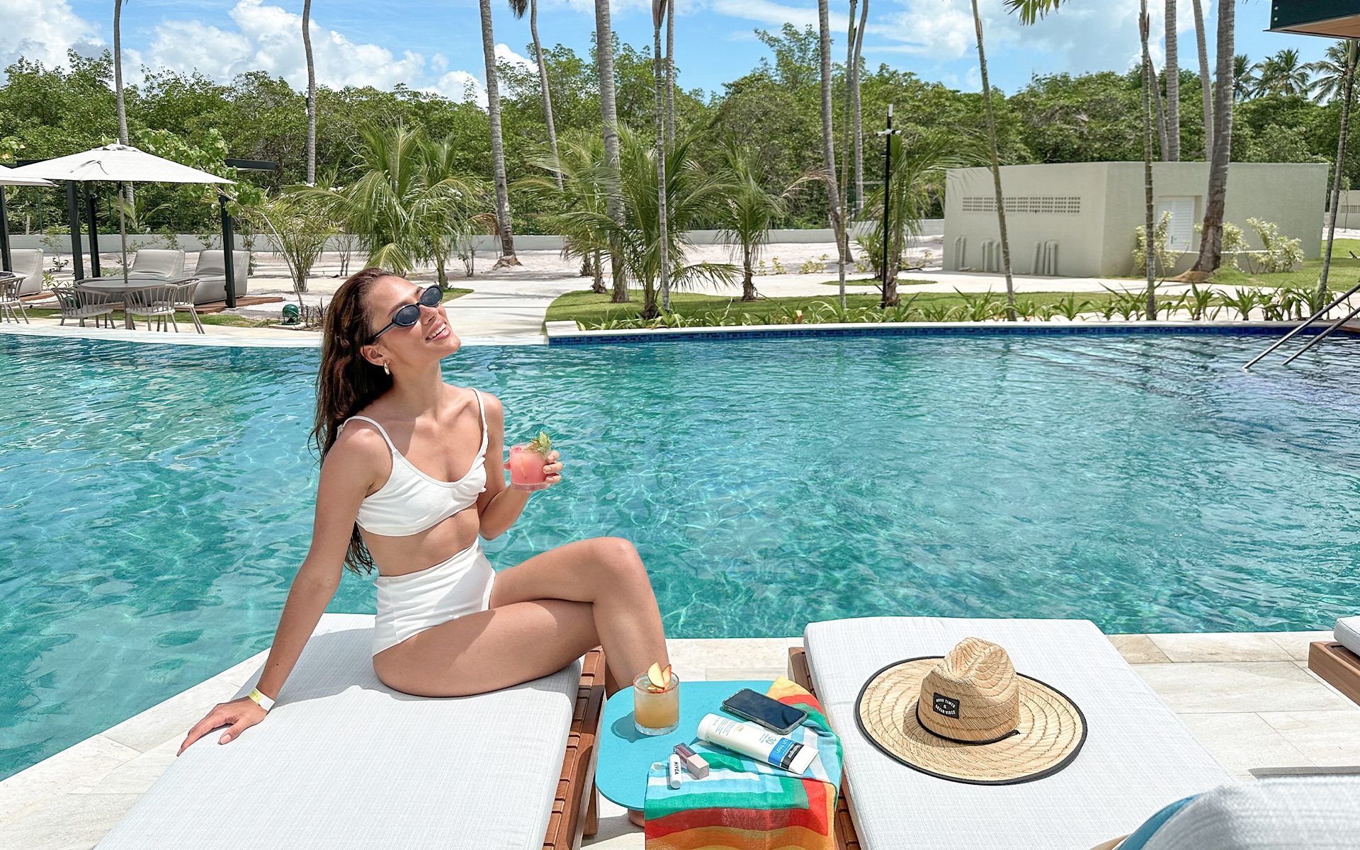 experiencia-japaratinga-lounge-resort Mulher sorridente relaxando em espreguiçadeira à beira da piscina na área das Gardens do Japaratinga Lounge Resort, segurando drink em dia ensolarado.