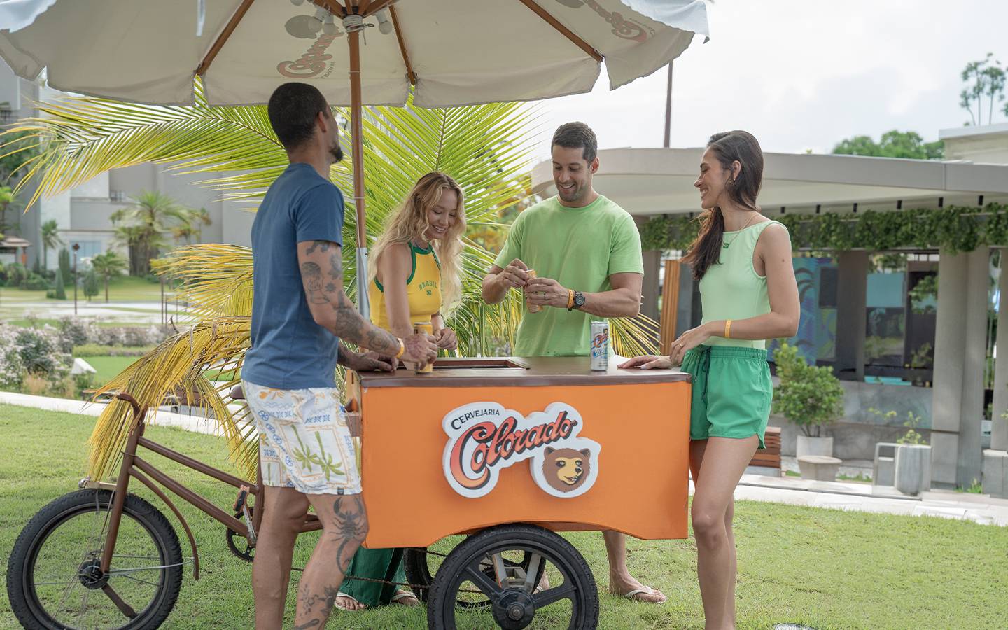 Grupo de amigos com roupas verdes e amarelas sorrindo ao redor de um carrinho de cerveja Colorado sob o ombrelone no Japaratinga Lounge Resort, em clima de torcida.