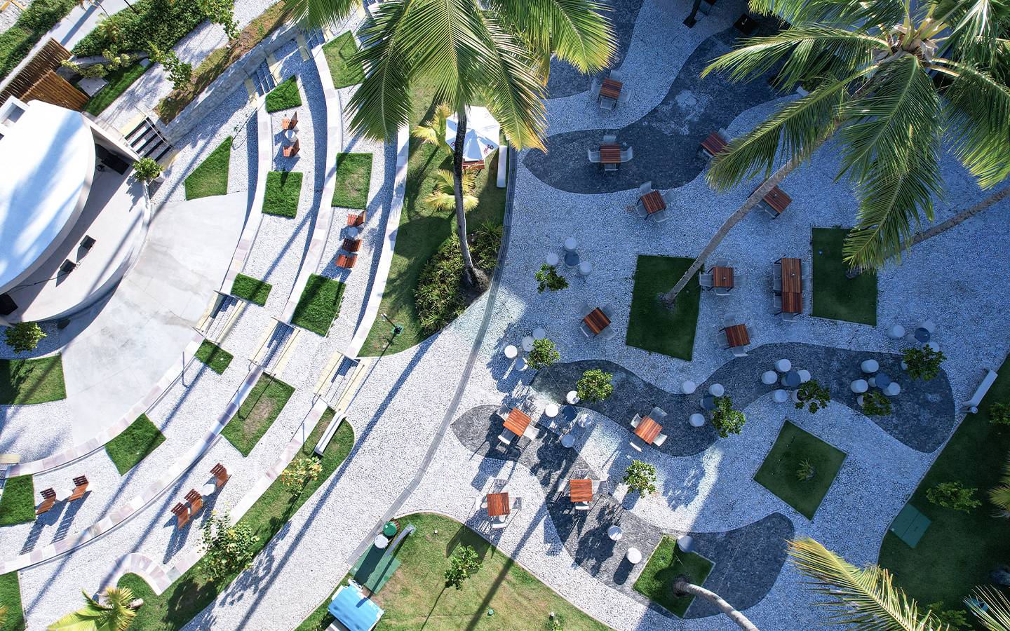 Vista aérea da Praça dos Coqueiros no Japaratinga Lounge Resort, com desenho de pedras no chão, muitos bancos e cadeiras pra sentar com mesas, áreas verdes com coqueiros, e no canto esquerdo um palco.
