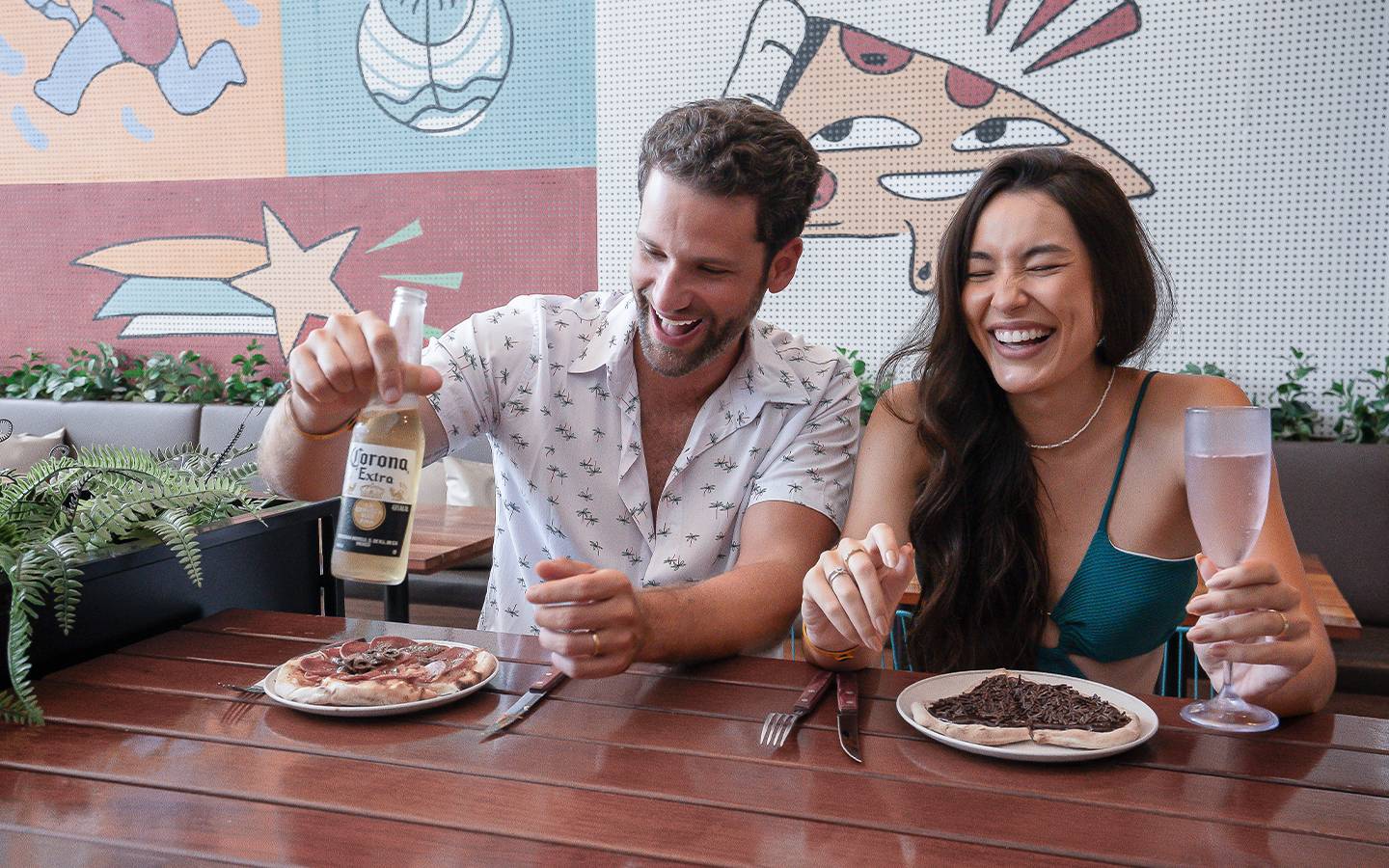 Casal comendo pizza na Forneria Amore, o homem come uma pizza salgada e está com cerveja Corona na mão e a mulher uma pizza de chocolate em formato de coração com taça na mão. Ao fundo uma parede decorada.