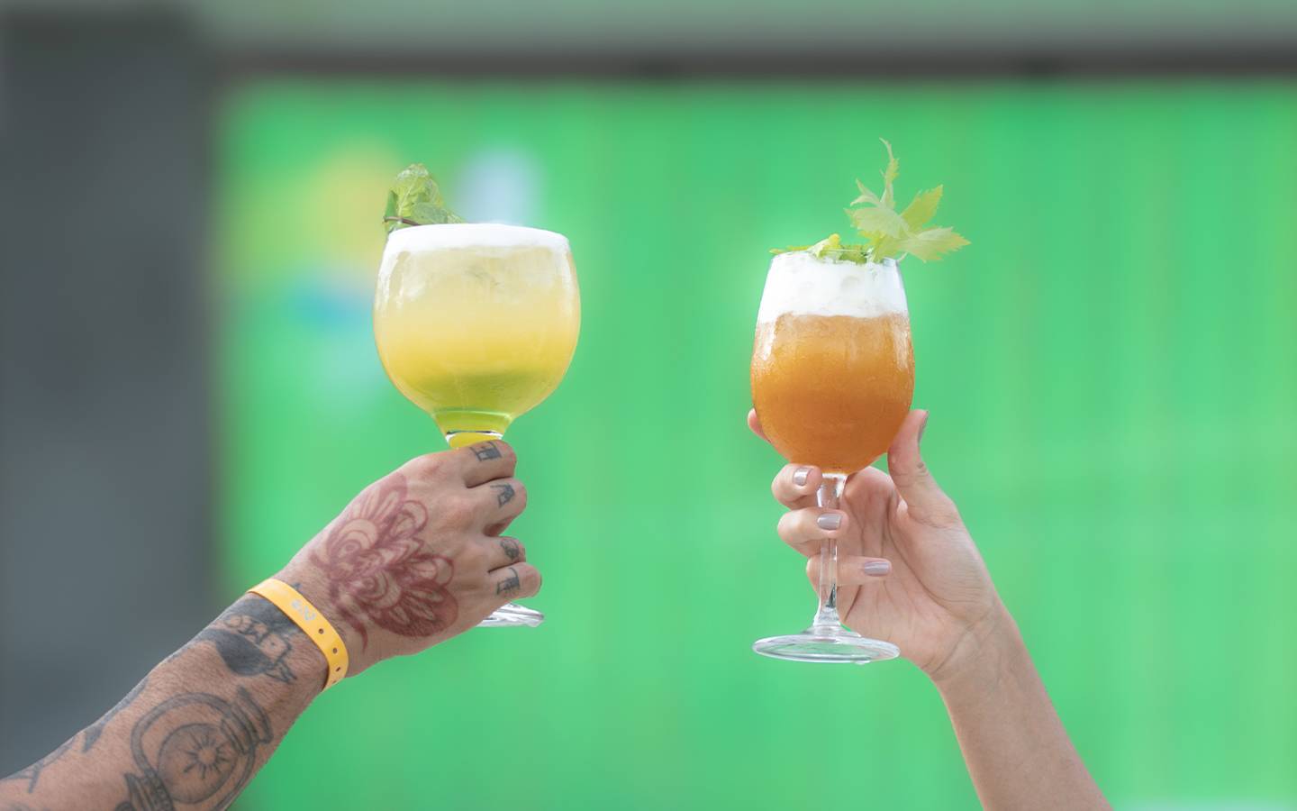 Dois bracos, um do lado esquerdo segundo um drink mais esverdeado com amarelo e um do lado direito com o drink mais alaranjado com espuma na parte de cima.
