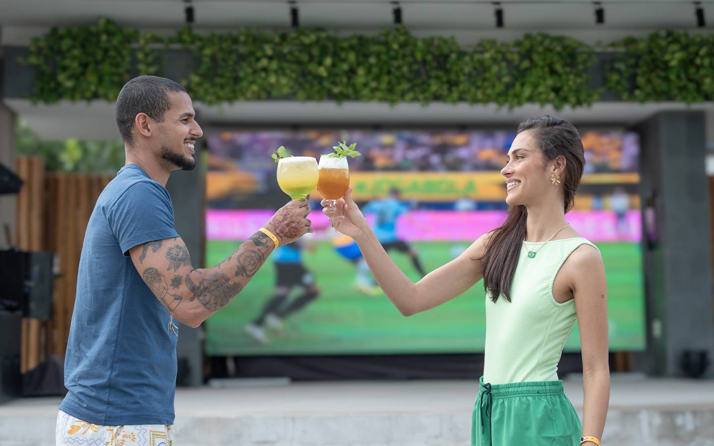 Casal jovem brinda com drinks coloridos em frente a um telão LED gigante exibindo um jogo de futebol (com jogadores em uniforme azul) no Japaratinga Lounge Resort, em clima de torcida e comemoração. Duas pessoas vestem roupas verdes, aludindo ao Brasil.