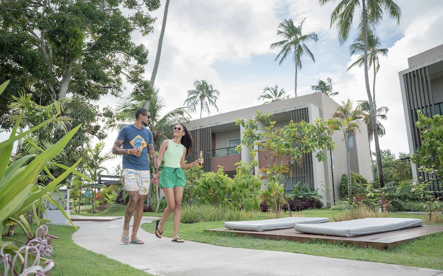 Casal com roupas verdes e amarelas sorrindo caminhando pelo resort com uma cerveja Corona na mão no Japaratinga Lounge Resort, em clima de torcida.