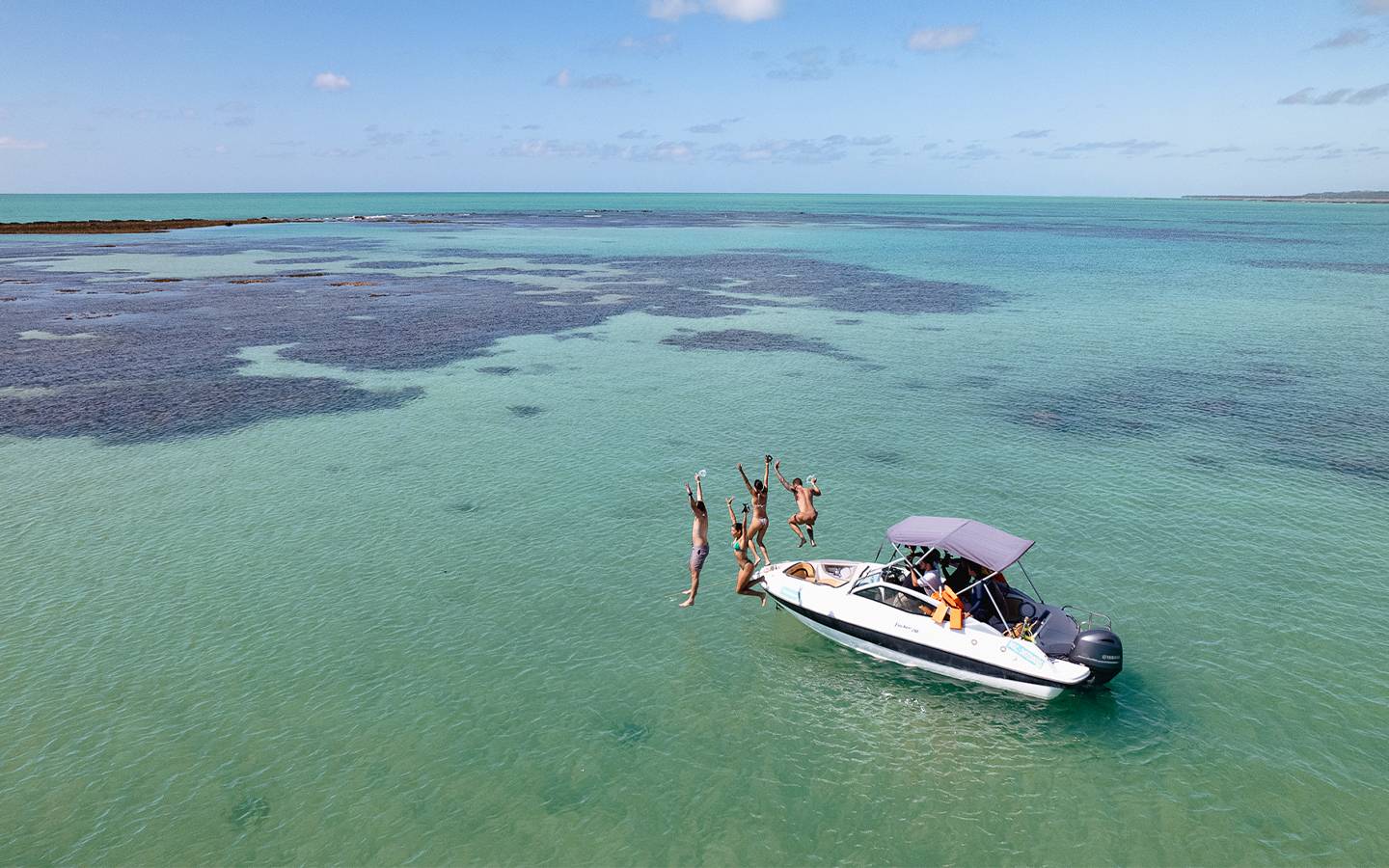Grupo de quatro amigos pulando do barco na Praia de Japaratinga. O mar está com maré baixa e céu sem nuvens, ao fundo o horizonte de mar e céu.