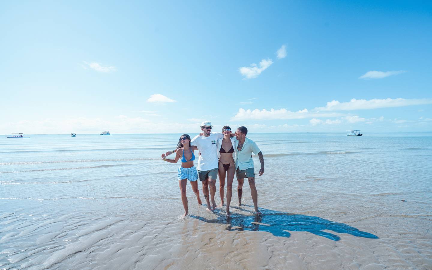 Grupo de quatro amigos abraçados na Praia de Japaratinga. Eles estão andando na beira da água, o mar está com maré baixa e céu sem nuvens, ao fundo do mar tem alguns barcos parados.