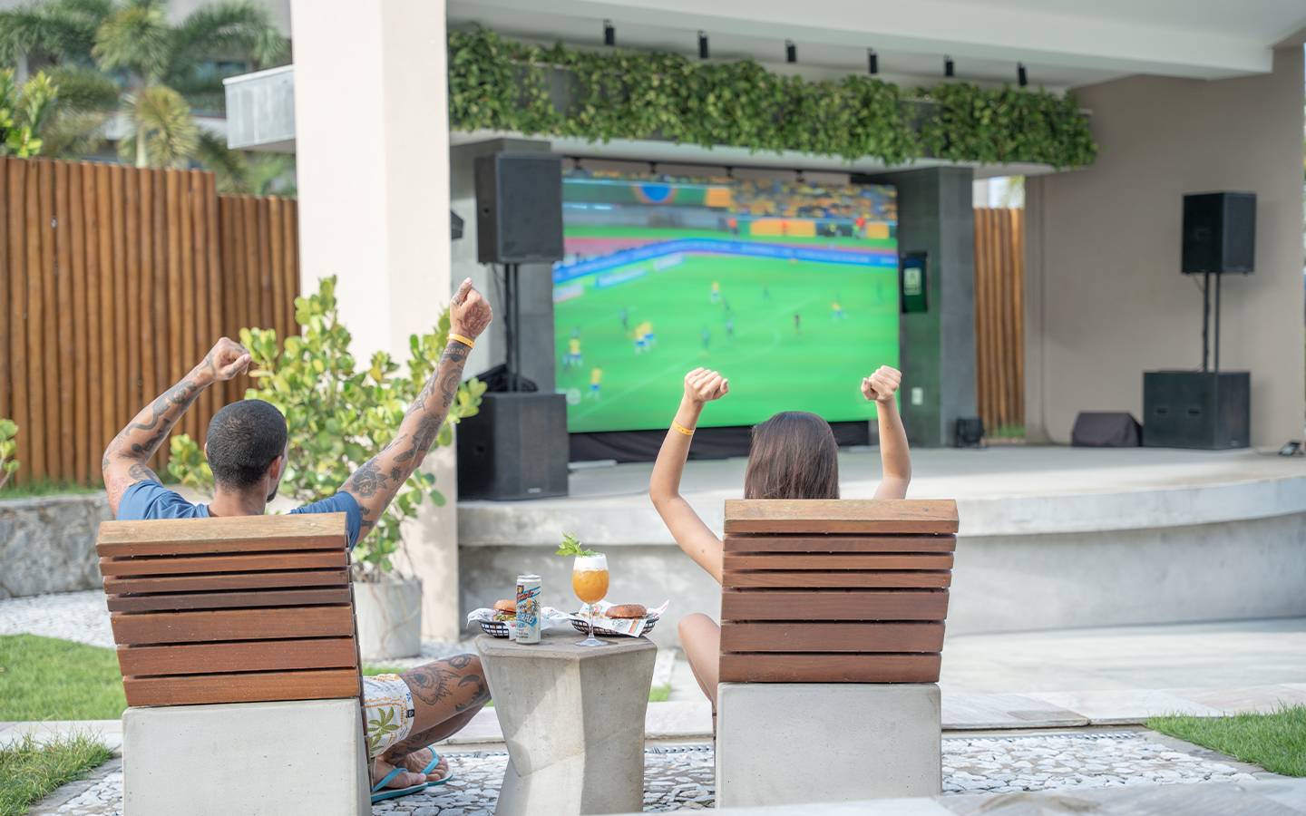 Casal sentado em espreguiçadeiras, com os braços erguidos em comemoração, em frente a um telão gigante exibindo partida de futebol no Japaratinga Lounge Resort, em clima de torcida.
