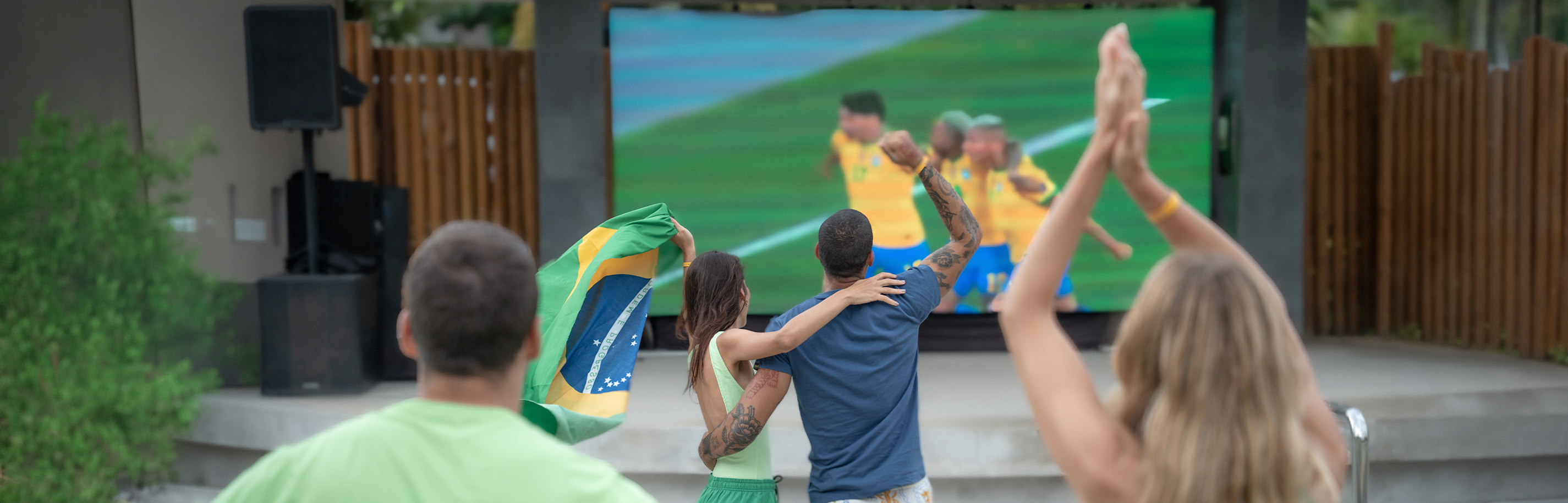 Casal jovem brinda com drinks coloridos em frente a um telão LED gigante exibindo um jogo de futebol (com jogadores em uniforme azul) no Japaratinga Lounge Resort, em clima de torcida e comemoração. Duas pessoas vestem roupas verdes, aludindo ao Brasil.