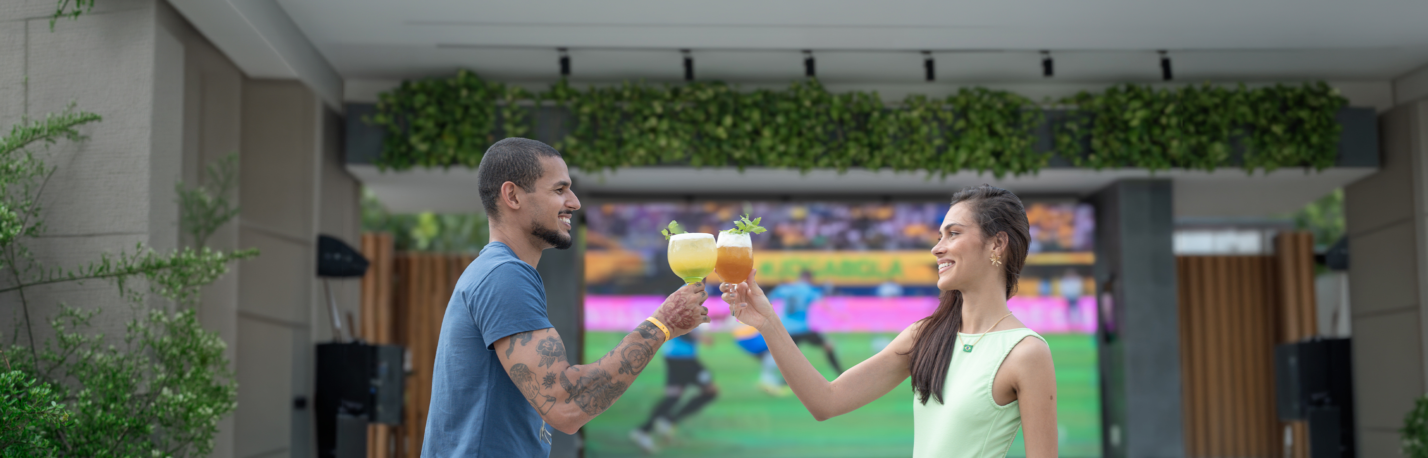 Casal jovem brinda com drinks coloridos em frente a um telão LED gigante exibindo um jogo de futebol (com jogadores em uniforme azul) no Japaratinga Lounge Resort, em clima de torcida e comemoração. Duas pessoas vestem roupas verdes, aludindo ao Brasil.