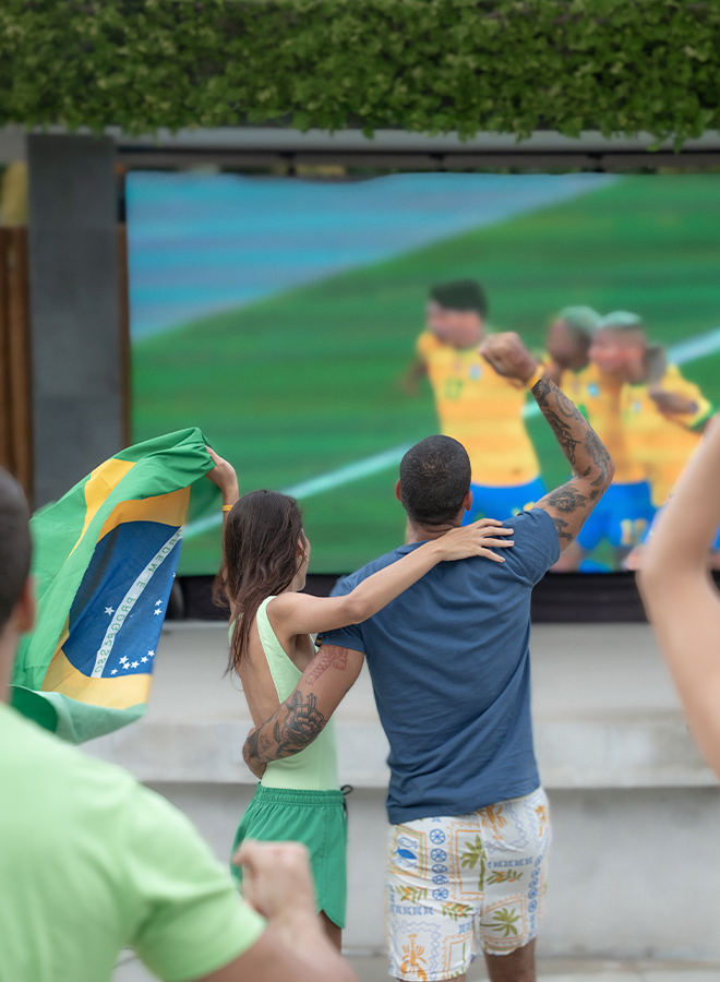Casal jovem brinda com drinks coloridos em frente a um telão LED gigante exibindo um jogo de futebol (com jogadores em uniforme azul) no Japaratinga Lounge Resort, em clima de torcida e comemoração. Duas pessoas vestem roupas verdes, aludindo ao Brasil.