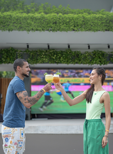 Casal jovem brinda com drinks coloridos em frente a um telão LED gigante exibindo um jogo de futebol (com jogadores em uniforme azul) no Japaratinga Lounge Resort, em clima de torcida e comemoração. Duas pessoas vestem roupas verdes, aludindo ao Brasil.
