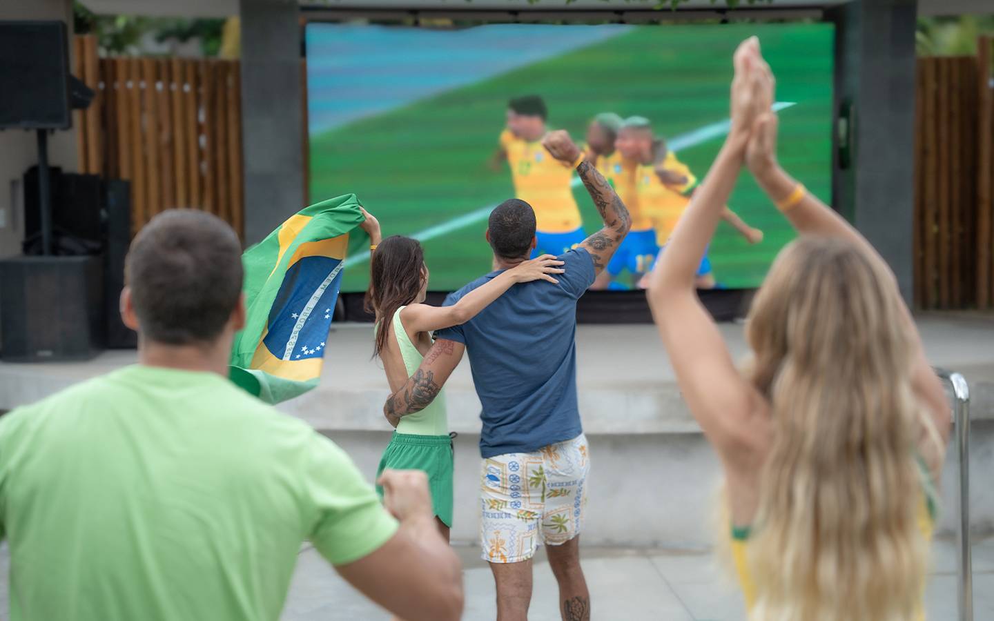 Quatro pessoas de costas, incluindo um casal abraçado com a bandeira do Brasil, torcem e comemoram em frente a um telão LED gigante que exibe um jogo de futebol (jogadores com uniformes amarelos e azuis) no Japaratinga Lounge Resort.