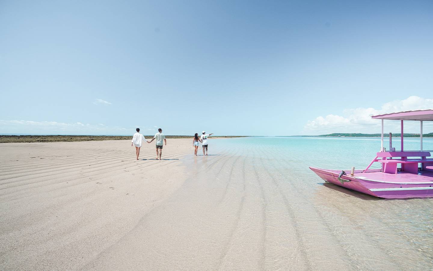 Vista aérea da Croa de São Bento, com bancos de areia, águas cristalinas azul-turquesa e jangada rosa ancorada. Ao fundo, um grupo de amigos caminha na faixa de areia.