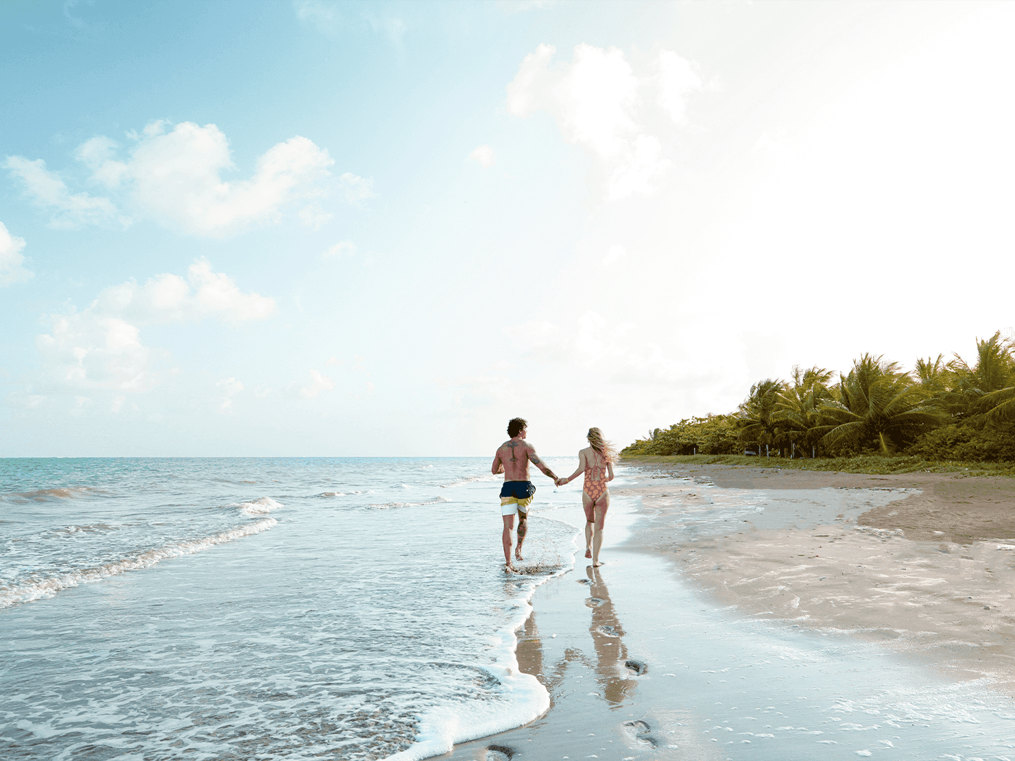 Casal caminhando de mãos dadas na beira do mar na Praia do Toque, com ondas suaves, areia clara, coqueiros ao fundo e céu azul com nuvens.