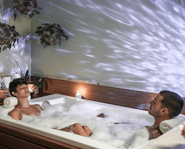 Casal feliz e sorrindo na cama do quarto, com a mulher deitada na cama e o homem sentado. Na cama, há uma bandeja com taças e velas.